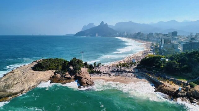 Drone flies over Pedra do Arpoador and Ipanema Beach, showcasing beautiful white sandy beach, blue ocean, palm trees, skyscrapers, and mountains in Rio de Janeiro