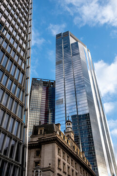 The image captures a cityscape with towering skyscrapers featuring modern glass facades reflecting the blue sky and clouds above, emphasizing urban architecture in London UK