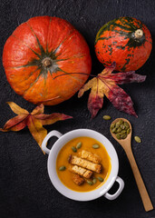 on a textured black background, top view, a white bowl of pumpkin soup garnished with pumpkin seeds and croutons. On the side, pumpkins and autumn leaves.