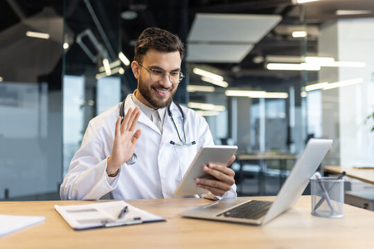 Smiling doctor wearing glasses and white coat engaged in virtual meeting on tablet. Modern technology enhances healthcare consultations for remote patients.