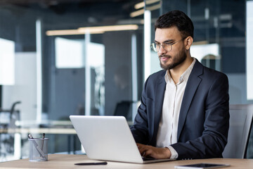 Businessman in modern office using laptop, focused on corporate task. Professional attire and...