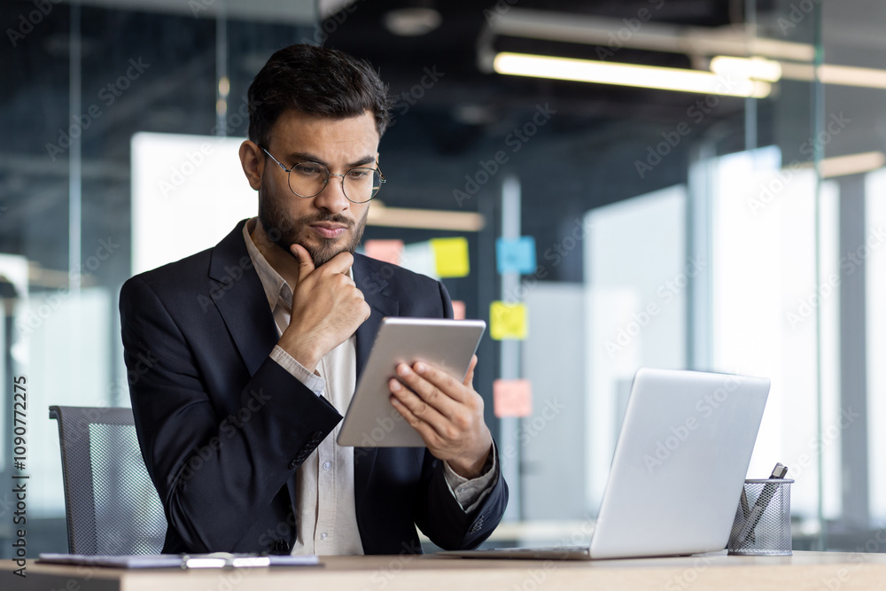 Wall mural Professional businessman in modern office uses tablet and laptop, displaying thoughtful expression. Concept of technology, analysis, business. Dressed in formal suit, engaged in workplace tasks.