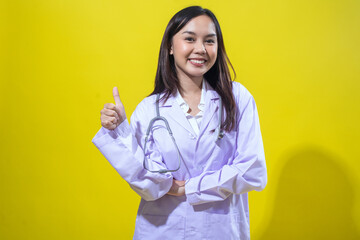 An Asian woman in a white doctor’s coat gives a cheerful thumbs-up while smiling confidently. She...