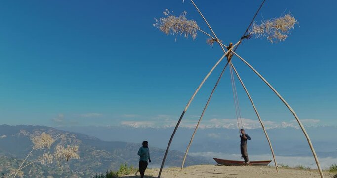 Drone shot captures two locals in Nepal swinging on a bamboo rope, a popular Dashain activity, with stunning mountain ranges, clear blue sky, and a festive, peaceful atmosphere