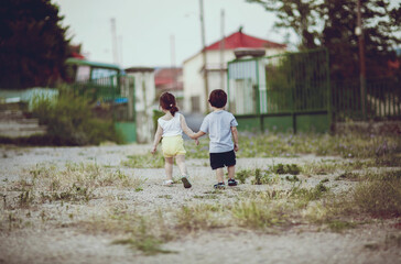 Two children stroll together along a gravel path, holding hands and enjoying their time outdoors. The backdrop includes homes and greenery, creating a peaceful atmosphere.