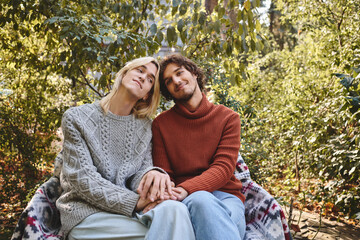 Fototapeta premium Two handsome young people sit closely together in a cozy embrace, sharing warmth and smiles amidst lush green surroundings. Their relaxed demeanor reflects love and connection.