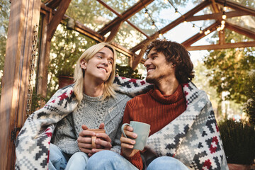 Two young men, smiling and enjoying each others company, cozy up under a colorful blanket, sipping...