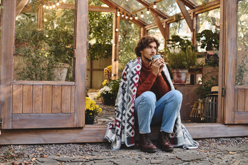 A young person relaxes outside a greenhouse, sipping a warm beverage. Surrounded by lush greenery...