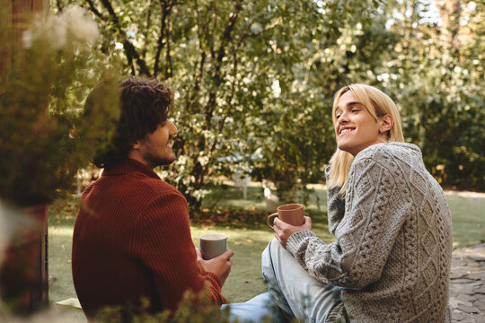 In a serene outdoor setting, Young loving gay couple with diverse styles relax together. With cups in hand and laughter in the air, they embody joy amidst lush greenery.