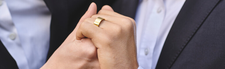 Fototapeta premium Two young and handsome men clasp hands, showcasing a beautiful golden ring that symbolizes their deep bond. Surrounded by warm sunlight, they share a tender moment filled with joy.
