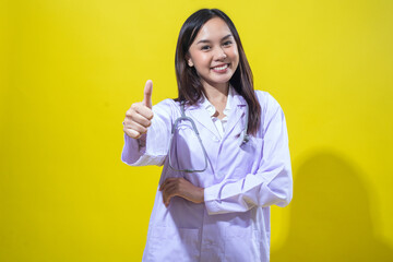 An Asian woman in a white doctor’s coat gives a cheerful thumbs-up while smiling confidently. She...