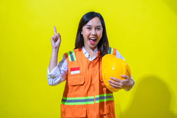 An Asian woman in an orange reflective safety vest holds a yellow helmet while pointing upward...