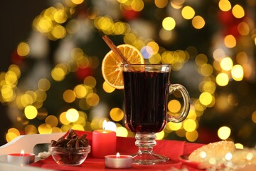 Aromatic mulled wine in glass cup, anise, cookies and burning candles on tray against blurred Christmas lights, bokeh effect