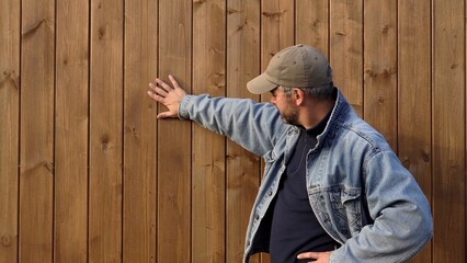 A man points at a wooden wall. Bearded. 40-45 years old. Wearing a denim jacket. Outdoors. Alone....