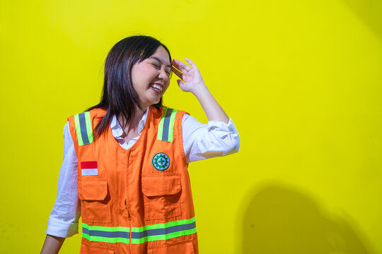 An Asian woman in an orange safety vest smiles brightly with her eyes closed, playfully shielding her face with her hand. She stands against a vibrant yellow background, exuding joy and positivity. - Powered by Adobe