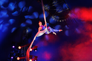 Aerialist performs a dangerous trick on a fabric high under dome of the circus under the spotlight.