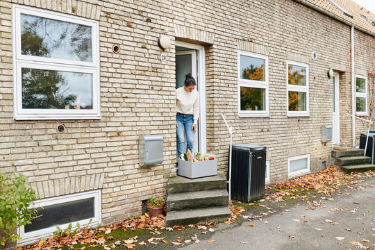 A woman stands in front of her front door, looking down at a grocery box that just has been delivered. The box is filled with organic vegetables and bread. The autumn leaves are lying by her house.
