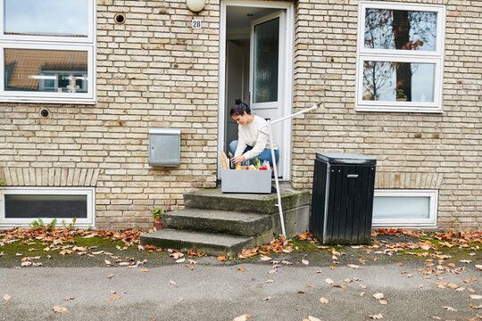A woman squats in front of her front door, looking through a grocery delivery box she just received. The box is filled with fresh organic vegetables and bread. The autumn leaves are lying by her house