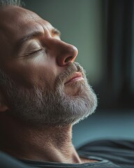 Obraz premium A middle-aged man with a gray beard, eyes closed, enjoying a moment of relaxation indoors.