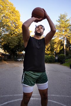 A basketball player prepares to take a precise shot, holding the ball overhead on an outdoor court surrounded by trees and illuminated by warm sunlight in a calm park setting