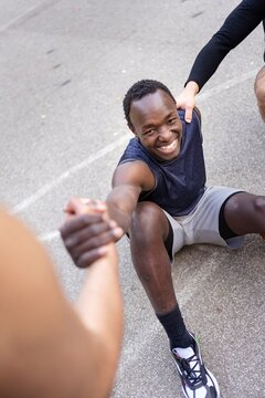 A smiling basketball player reaches out to be helped up by teammates during a game on an outdoor court surrounded by trees and soft sunlight