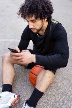 A basketball player sitting on the court holding a phone, smiling and enjoying a break on a sunny day surrounded by trees and a calm park atmosphere