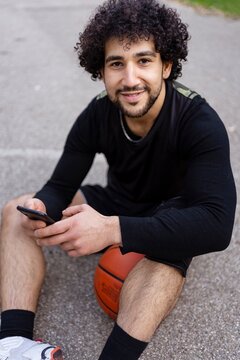 A basketball player sitting on the court holding a phone, smiling and enjoying a break on a sunny day surrounded by trees and a calm park atmosphere