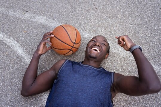 A joyful basketball player lying on the court holding the ball and smiling brightly under soft sunlight on a peaceful park court surrounded by greenery
