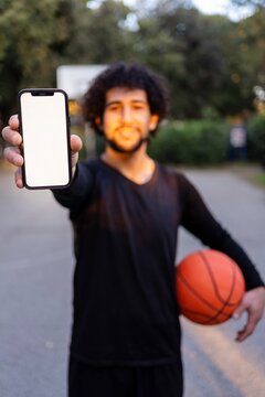 A smiling basketball player holding his phone and ball, posing on an outdoor court under warm sunlight surrounded by park greenery