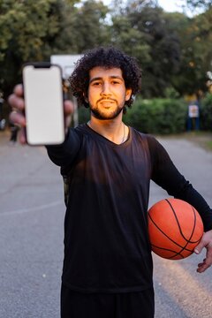 A smiling basketball player holding his phone and ball, posing on an outdoor court under warm sunlight surrounded by park greenery