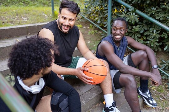 Three basketball players sit on outdoor steps, smiling and talking while holding basketballs, surrounded by lush greenery in a relaxed park environment