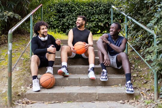 Three basketball players relax on a set of outdoor steps, laughing and chatting while holding basketballs, surrounded by greenery in a peaceful park setting