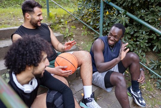 Three basketball players sit on outdoor steps, smiling and talking while holding basketballs, surrounded by lush greenery in a relaxed park environment