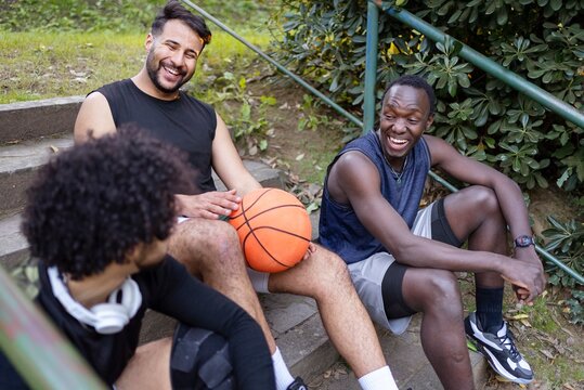 Three basketball players sit on outdoor steps, smiling and talking while holding basketballs, surrounded by lush greenery in a relaxed park environment
