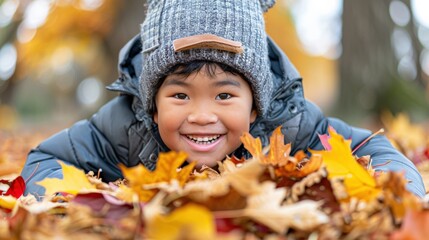 Fototapeta premium Smiling Asian boy wearing gray winter hat dark jacket lying on colorful autumn leaves in park, background blurred with warm golden tones, expressing joy, excitement, seasonal outdoor activity.