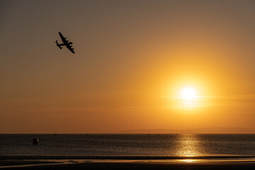 The sun sets over Ayr beach, and the silhouette of a flying bomber is visible in the sky. Which...