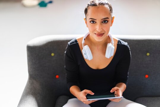 A woman dressed in ballet attire sits on a gray sofa with white headphones around her neck, holding a tablet and looking forward with a calm expression in a bright room - Powered by Adobe