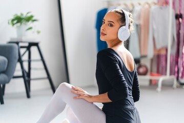A ballerina wearing white headphones and a black top sits on the studio floor, gazing confidently with clothing racks and greenery decorating the bright and airy studio background
