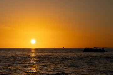 Obraz premium Golden sunset over the ocean with a silhouetted boat and a flying bird, reflecting warm light across the calm water.