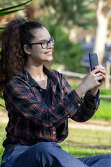 Young white girl in casual clothes takes a photo with her mobile phone in a green park while smiling