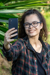 Young white girl in casual clothes takes a selfie with her mobile phone in a green park while smiling