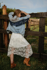 A young girl poses in a boho wedding dress, cowboy boots, a cowboy hat, and a denim jacket.