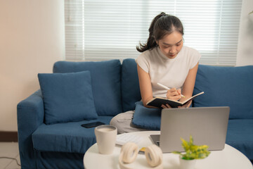 Comfortable young asian woman sitting on a blue sofa in her living room, working from home and...