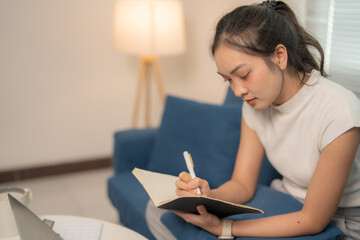 Fototapeta premium Concentrated young asian woman writing notes in her notebook while sitting on a comfortable blue sofa in her living room, working or studying from home