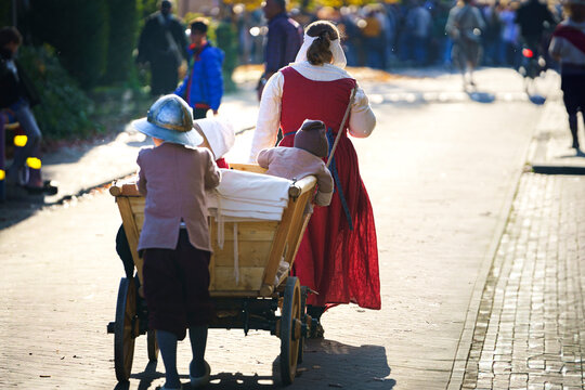 Young child pushes a handcart while a woman in medieval clothing drags a poor young family through the streets of a historical Dutch town