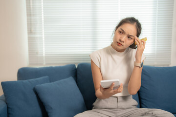 Young asian woman calculating domestic bills, touching her head with a credit card and looking...