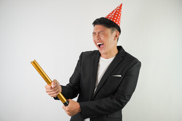 Excited young asian man wearing red party hat, white shirt and black suit is holding confetti cannon, isolated over white background. Concept for Christmas Holiday and New Year Party