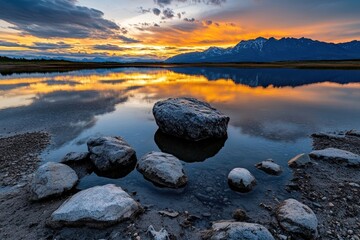A stunning landscape featuring tranquil water reflecting a vibrant sunset and majestic mountains, with rocks in the foreground.