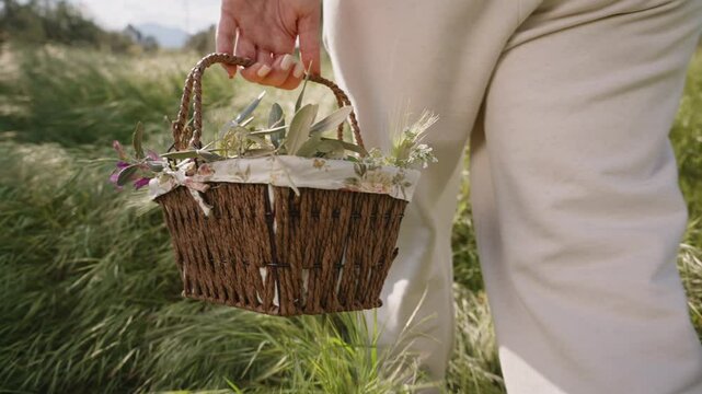 A Woven Basket Filled with Vibrant Flowers and Easter Eggs is Carried by a Woman in White Pants, Walking Through a Sunlit Field. Slow motion.