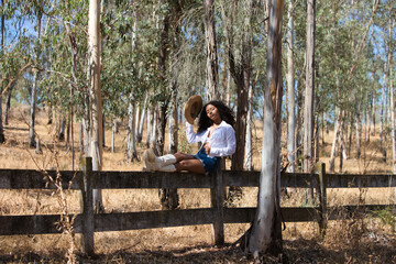 Fototapeta premium woman, young, beautiful, Latin, brunette with curly hair dressed in white shirt, denim skirt, white boots and hat, sitting with her legs stretched out on the fence of the eucalyptus forest.